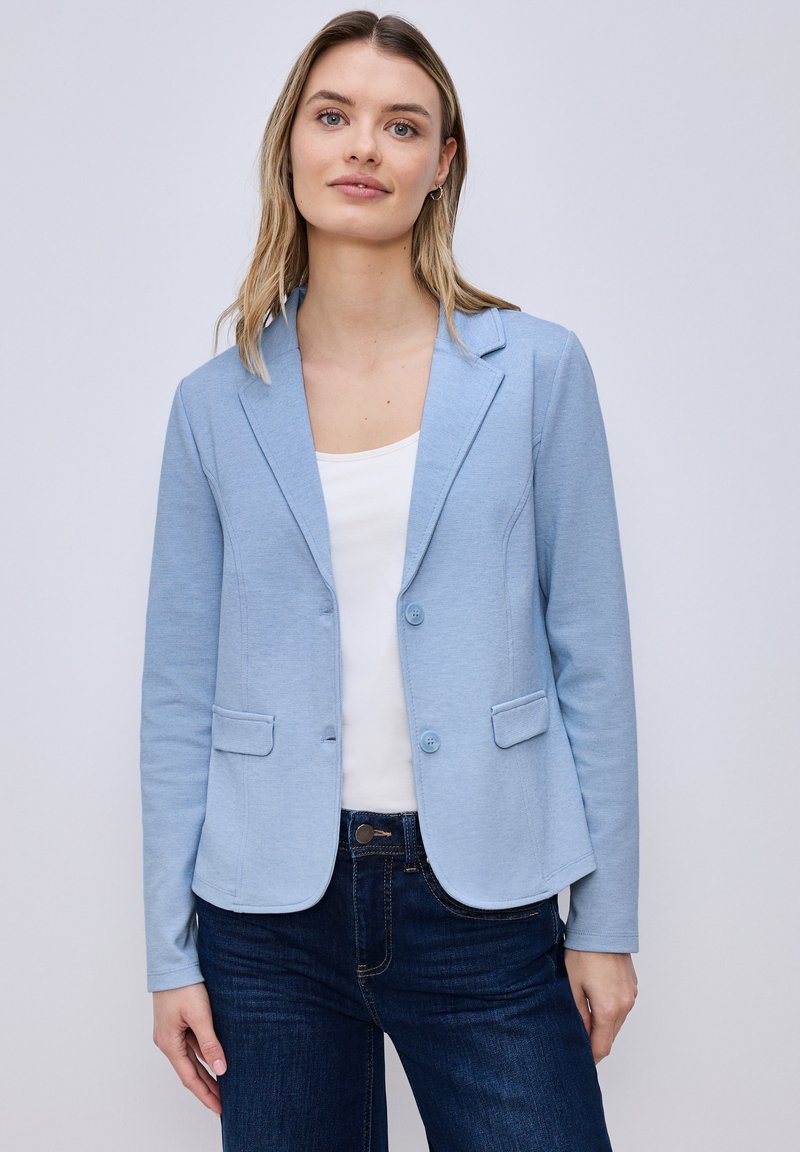 Jeune femme portant un blazer bleu clair sur un haut blanc et un jean foncé, debout devant un fond clair uni.
