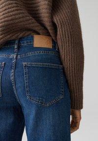 Jeans in denim blu con un maglione a maglia marrone texturizzato che copre la parte superiore. Presenta un'etichetta in pelle visibile con logo sopra le tasche posteriori.