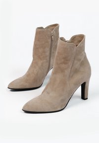 Beżowe zamszowe botki za kostkę z wysmuklonymi noskami, suwakami po bokach i średniej wysokości cienkimi obcasami na białym tle.