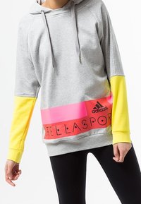 Sweat à capuche gris à coupe large, manches jaunes et bande horizontale rose avec "STELLASPORT" et le logo Adidas. Tissu en mélange de coton.