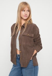 Młoda kobieta z długimi blond włosami, ubrana w brązową, guzikiem zapinaną kurtkę z korduroju na jasnoszarym topie i niebieskich dżinsach, z rękami w kieszeniach.