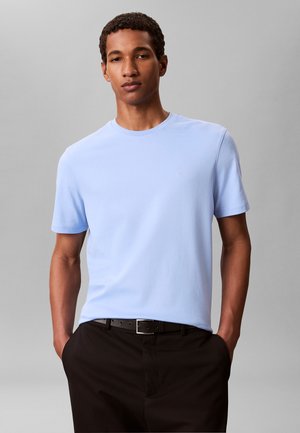 Mann trägt ein hellblaues kurzärmeliges T-Shirt und schwarze Hose, Hände in den Taschen, steht vor einem schlichten grauen Hintergrund.