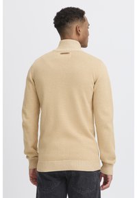 Suéter de punto beige con un cuello alto y puños acanalados. Textura suave con un ligero brillo, con una pequeña etiqueta en la parte posterior del cuello.
