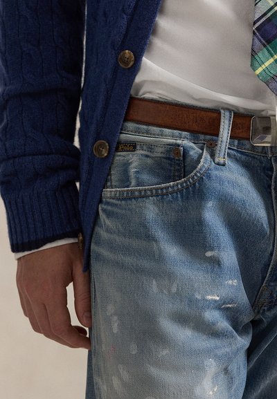 Personne portant un cardigan en maille torsadée bleu, une chemise blanche, une cravate à carreaux, une ceinture marron et un jean délavé bleu clair avec une étiquette "Polo" sur la poche.