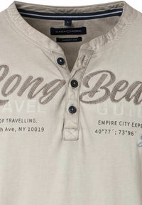 Beżowa koszula z krótkim rękawem i dekoltem henley, z dwiema guzikami oraz haftowanym napisem. Charakteryzuje się miękką fakturą i niebieskim akcentem na kołnierzyku.