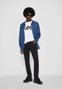 Persona alta con gafas de sol negras, camiseta blanca de Lee, camisa de mezclilla azul abierta, pantalones negros ajustados y botas negras contra un fondo blanco.