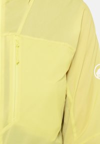 Lichtgele jas met verticale ritszak op de borst en een wit circulair logo op de bovenarm.