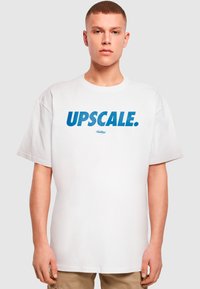 Hvid oversized t-shirt med fed blå tekst "UPSCAL" på forsiden. Lavet af blødt bomuld, den har en rund halsudskæring og korte ærmer.