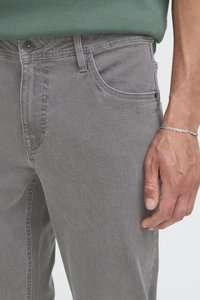 Mees, kes kannab halli denimpükst ja hõbedast ketist käevõru, rohelist särk vöökohas sissetõmmatuna.