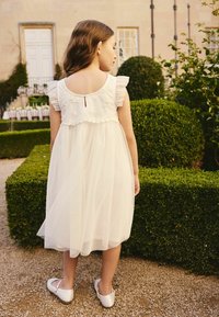 Witte tulejurk met geborduurde accenten, geruite mouwen en een rug met een sleutelgat en knoopsluiting. Gezien van achteren, gedragen met eenvoudige witte schoenen.