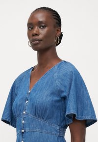 Vestido de mezclilla con escote en V, mangas cortas con volantes y detalles de botones. Cuenta con una cintura fruncida y una textura suave en un color azul sólido.