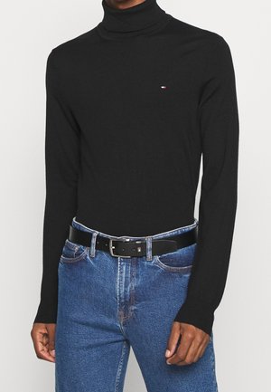 Uomo che indossa un maglione nero a collo alto e jeans blu con una cintura nera con fibbia argentata.