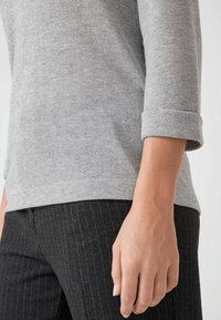 Arm und Oberkörper einer Person, die einen hochgekrempelten grauen Pullover und schwarze Nadelstreifenhosen trägt, vor einem schlichten Hintergrund.
