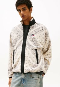 Giacca in pile con motivo paisley in crema e grigio, dotata di zip nera e colletto, e tasche laterali. Emblema sul lato sinistro.