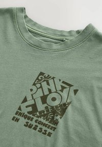 Grünes Baumwoll-T-Shirt mit einer strukturierten Grafik in Dunkelbraun, die den Text "Pink Floyd" mit dekorativen Elementen und Veranstaltungsdetails zeigt.