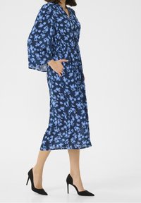 Mekko, jossa on laivastonsinisiä kukkia, pitkät ja levenevät hihat, vyötärö kireänä ja taskut. Mustat korkokengät täydentävät asua.