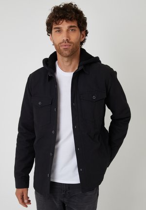 Czarna kurtka z kapturem, posiadająca miękką polarową podszewkę. Dwie kieszenie z przodu, zapięcia na guziki i długie rękawy. Nosi się ją na białym t-shircie.