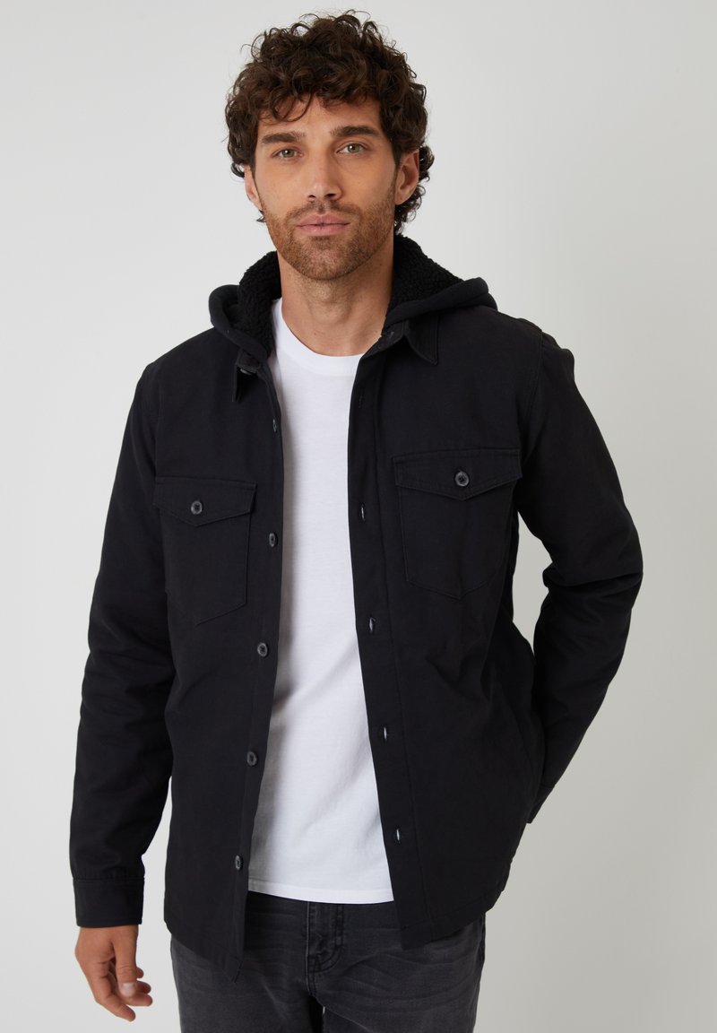 Czarna kurtka z kapturem, posiadająca miękką polarową podszewkę. Dwie kieszenie z przodu, zapięcia na guziki i długie rękawy. Nosi się ją na białym t-shircie.