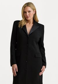 Melnā pielāgotā mēteli ar garām piedurknēm, ar iespiestu atlokiem, piecu pogu aizdari, divām priekšējām kabatām un nedaudz teksturētu audumu.