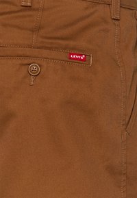 Hnědé bavlněné kalhoty s viditelnou šitým kapsou, vybavené kulatým knoflíkem a červenou visačkou s logem Levi's na boku.