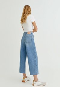 Femme aux cheveux blonds ondulés portant un haut blanc à manches courtes, un jean large bleu clair et des baskets blanches, se tenant devant un fond uni.