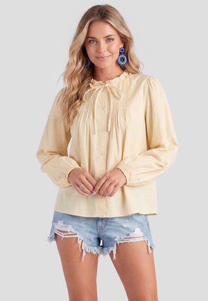 Frau trägt eine hellgelbe langärmelige Bluse mit Knöpfen vorne und ausgefranste blaue Jeansshorts, steht mit verschränkten Händen.