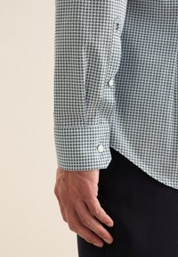 Camicia a maniche lunghe con bottoni di colore verde chiaro e un piccolo motivo geometrico. Il tessuto ha una consistenza liscia e presenta bottoni bianchi nei polsini.