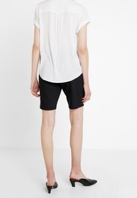 Biała bluzka z krótkim rękawem i podwiniętymi mankietami, zestawiona z czarnymi szortami. Czarne mule dopełniają zestaw. Prosty design, gładka tekstura.