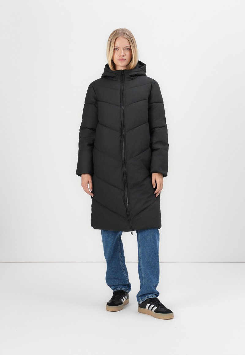 Doudoune noire avec capuche, dotée de panneaux matelassés horizontaux, d'une fermeture éclair frontale et de poches. Portée sur un jean bleu et des sneakers noires.