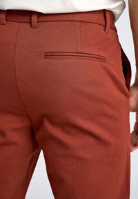 Pantaloni rossi con texture, caratterizzati da una tasca posteriore con apertura stretta e passanti per la cintura, posizionati sopra una mano appoggiata sull'anca.