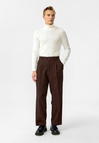 Licht crème coltrui en donkerbruin gestreepte broek. Schoenen zijn zwart en de broek heeft een knoopsluiting en plooien.