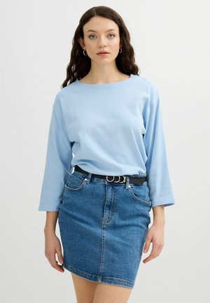 Joven mujer con cabello largo y oscuro que lleva una camisa de manga larga azul claro, falda vaquera, cinturón negro con anillos plateados y pendientes de aro dorados.