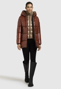 Bruine pufferjas met een capuchon, met een boxy, gestikt ontwerp. Binnenste lichtbeige jas, zwarte leggings en kniehoge zwarte laarzen. Effen achtergrond.