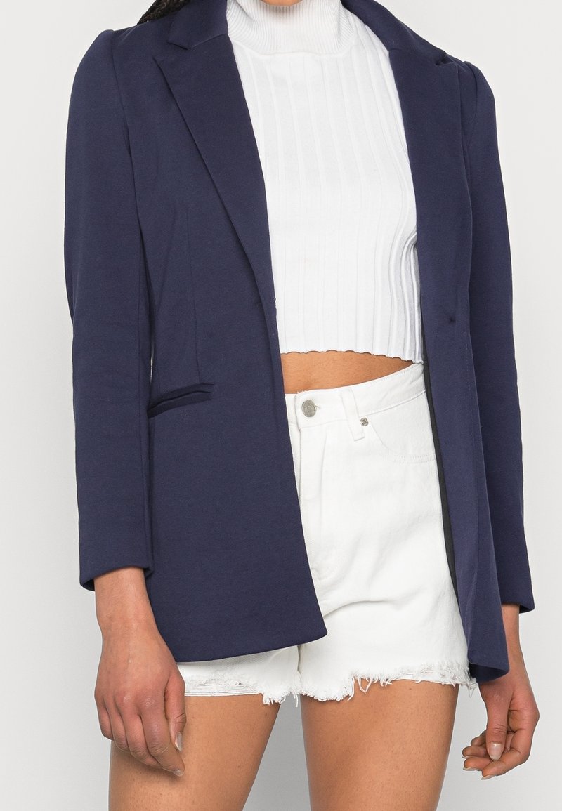 Mörkblå kavaj med långa ärmar, enkelknäppning och detaljer med framfickor, bärs över en vit ribbad turtleneck och vita shorts.