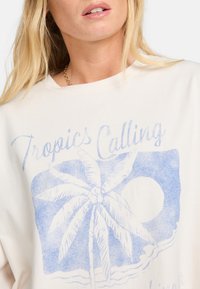 Lysebeige bomulds-T-shirt med et blåt grafisk motiv af et palmetræ og en sol, med teksten "Tropics Calling" ovenfor designet.