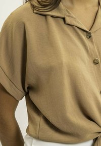 Chemise brune à manches courtes avec col, dotée de deux boutons et d'un tissu doux et texturé. Le détail en nœud à la taille ajoute un élément de design.
