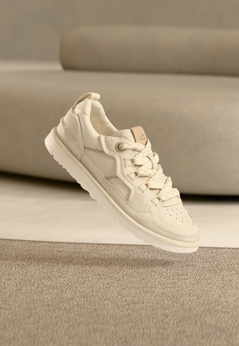 Baskets couleur crème avec une tige texturée, un bout perforé et une fermeture à lacets. Elles présentent un logo discret et une semelle blanche rembourrée.