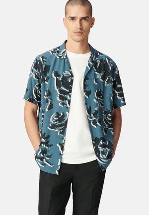 Kurzarmhemd in Teal mit floralen Mustern in Schwarz und Weiß. Verfügt über ein Knopfdesign und eine entspannte Passform. Einfaches weißes Unterhemd sichtbar.