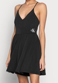 Zwarte mouwloze jurk van zachte stof, met een diepe V-hals, een uitlopende rok en een accent van het Calvin Klein-logo op de taille.