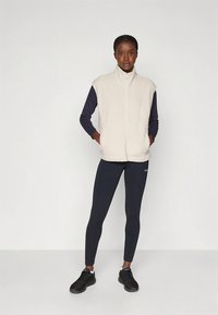 Off-white-värinen fleece-liivi korkealla kauluksella, edessä vetoketju, yhdistetty laivastonsiniseen pitkähihaiseen paitaan ja mustiin leggingsseihin. Mustat urheilukengät.