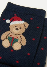 Marineblå sokker med en strikket teddybjørn iført en rød juletagd, der holder en gave med en rød sløjfe, omgivet af røde prikker.