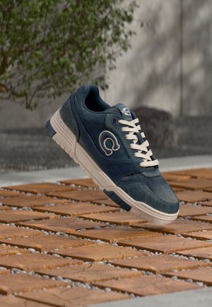 Dark blue sneaker with white laces and logo, suspended above a wooden tile surface outdoors with blurred plants and wall background.