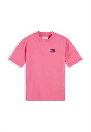 Lächelndes Kind mit lockigem Haar, das ein pinkes T-Shirt, schwarze Hose und weiße Sneakers trägt, steht vor einem weißen Hintergrund.