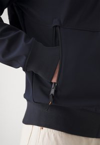 Ręka osoby w częściowo zapiętej na zamek kieszeni czarnej kurtki z prążkowanym mankietem, ubrana w jasne spodnie.