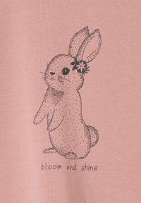 Illustration eines sitzenden Kaninchens mit einer Blume hinter dem Ohr auf einem rosa Hintergrund, mit dem Text "blühen und strahlen" darunter.