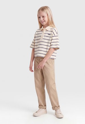 Usmiata mladá dievčina s dlhými blond vlasmi, oblečená v krémovo-modro pruhovanom polotriku, béžových nohaviciach a béžových teniskách, stojaca na bielom pozadí.