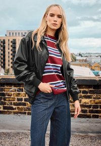 Giacca bomber nera in pelle con polsini a coste, abbinata a un maglione a righe bordeaux e jeans blu a gamba larga. Sfondo urbano.