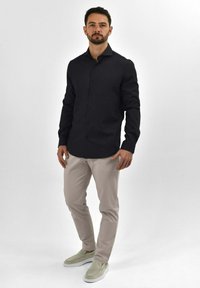 Donker, knooploze shirt met een nette kraag; lichtbeige slim-fit broek; casual instapschoenen van een zachte stof.