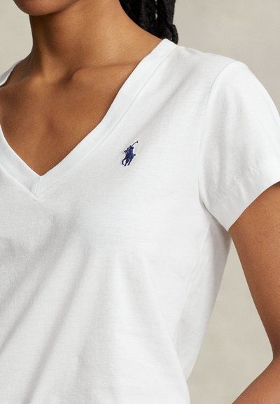 Kvinna iklädd en vit V-ringad T-shirt med en liten broderad polo-spelare i marinblått på vänster bröst.