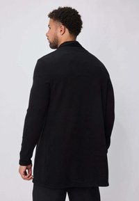 Long gilet noir en tissu doux, avec une texture lisse, devant ouvert et manches longues, arrivant sous les hanches.